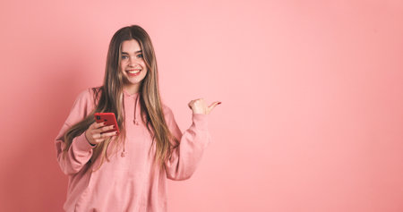 Happy woman holding smartphone and gesturing towards empty space on pink backgroundの写真素材