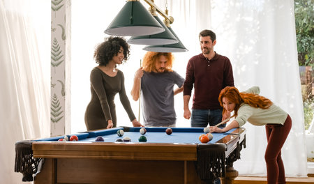 Woman aiming cue stick on billiard ball while playing pool with diverse friends at homeの写真素材