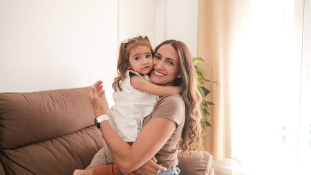 Smiling woman hugging cute daughter on sofa at homeの写真素材