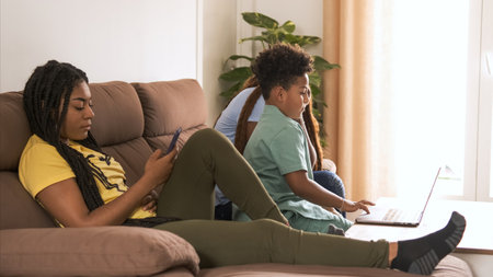 Woman using smartphone while brother and mother using laptop at homeの写真素材
