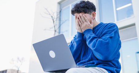 Stressed student covering face with hands while working on laptop outside universityの写真素材