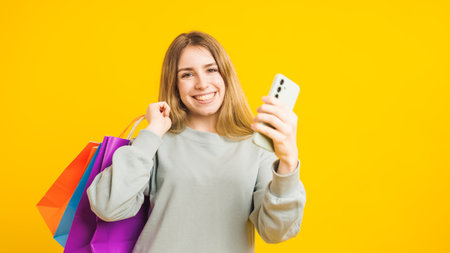 Happy shopper using smartphone and holding shopping bags on yellow backgroundの写真素材