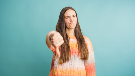 Young woman showing thumbs down gesture expressing disapproval on turquoise backgroundの写真素材
