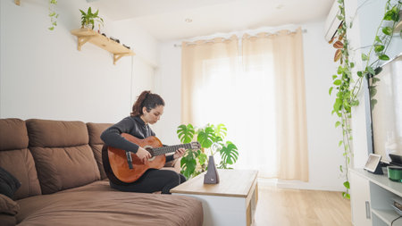 Young woman playing acoustic guitar at home during online lessonの写真素材