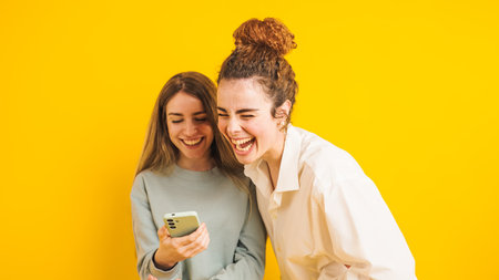 Two young women laughing while looking at smartphone on yellow backgroundの写真素材