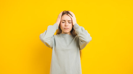 Young woman suffering from headache holding head with hands on yellow backgroundの写真素材