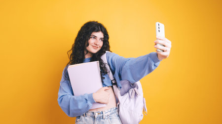 Smiling student taking selfie holding notebook and backpack on yellow background