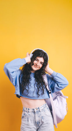 Young student listening music with wireless headphones and backpack on yellow backgroundの写真素材