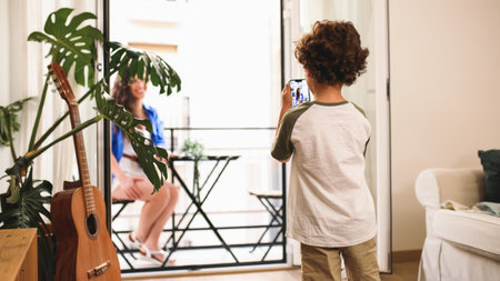 Child taking photo of mother relaxing on balcony with smartphoneの写真素材