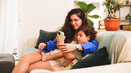 Mother and son watching smartphone sitting on sofa at homeの写真素材