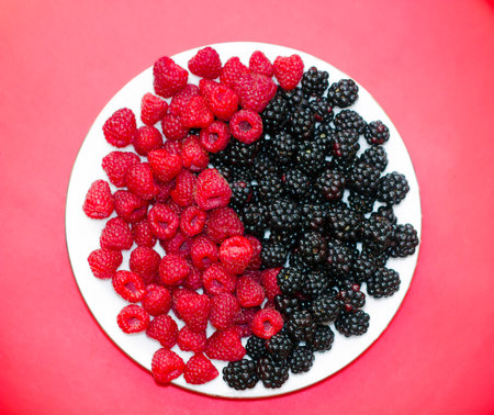Fresh raspberries and blackberries on plate lined with symbol of yin and yang on red backgroundの写真素材