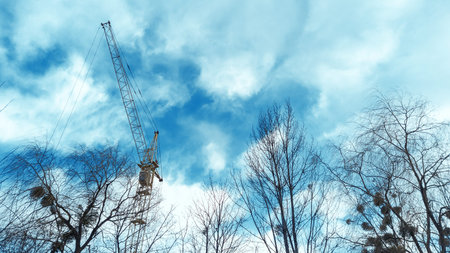 Tower crane by blue cloudy sky and branch of trees. Yellow high construction crane building house