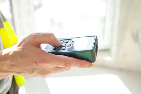 Laser rangefinder in man hand. Engineer in protective yellow vest, hold laser distance meter. Measuring device in house. Worker using electronic range finder on building, measure working conceptの写真素材