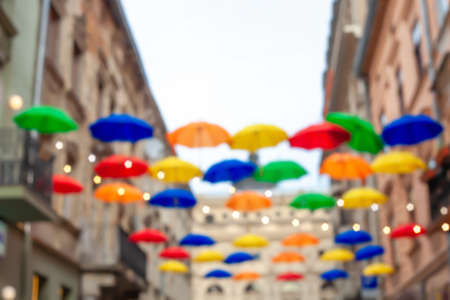 Blurred background with multicolored open umbrellas hanging on city street. Blurry lot of flying parasol and houses. Blue, orange, green, yellow and red sunshade in summer town abstract backdropの写真素材