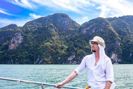 Portrait of young man on blue sea and green island background, summer tropical vacations. Man wearing white shirt resting on yachtの写真素材