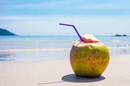 Green coconut water drink with drinking straw on tropical sandy beach by blue sea. Cold beverage in sand by turquoise ocean at sun light dayの写真素材