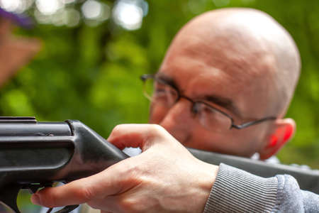 Young man shooting a gun. Man aiming with shotgun on shooting range close up. Hunter in summer green forest closeup, soft selective focus on human handの写真素材