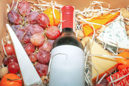 Cardboard gift box with red wine and fruit closeup. Present box with cheese, grapes, tangerines, and wine bottle mock up. Gourmet christmas hamper top viewの写真素材