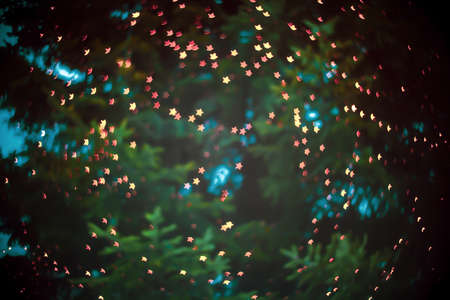 Blurred background with small stars on christmas tree. Abstract defocused shiny bright backdrop. Festive star bokeh effect by night dark blue sky. Xmas vivid starry glow ornament on green pine treeの写真素材