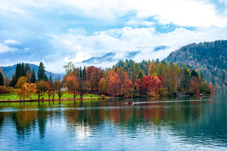 Autumn lake Bled, Julian Alps mountain, boats at distance in Slovenia. Serenity landscape over Blejsko jezero, mounts in cloud, floating wooden boat on reflection water in Carniolan region Slovenijaの写真素材