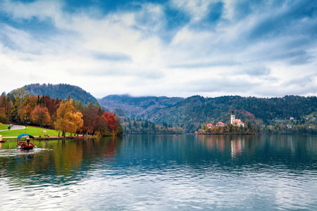 Autumn sunset lake Bled, church of Assumption of Mary Pilgrimage on island, swimming boat in Slovenia. Fall landscape over Blejsko jezero, mountains in cloud, pletna on water in Carniolan Slovenijaの写真素材