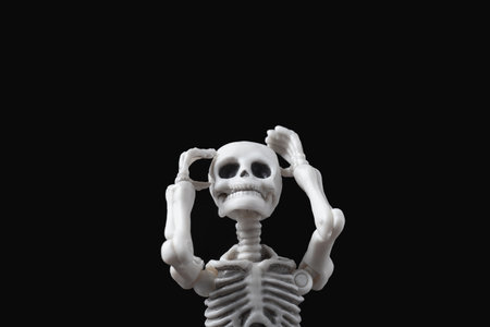 Skeleton figurine hold its head by black background. Desperate figure on dark backdrop. Stressed and sad doll, mental problems, feeling bad, depressed, disappointment, hopeless emotions conceptの写真素材