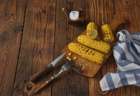 cobs of corn on a wooden table with cutlery, salt shaker and cloth napkin, top view