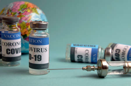 a syringe with a drop of medicine on a needle, vials with a vaccine against the background of the globe in disfocus on a light blue-green backgroundの写真素材