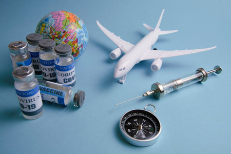 a syringe with a drop of medicine on a needle, vials with a vaccine on the background of the globe, an airplane and a compass, a concept, on a light blue-green backgroundの写真素材