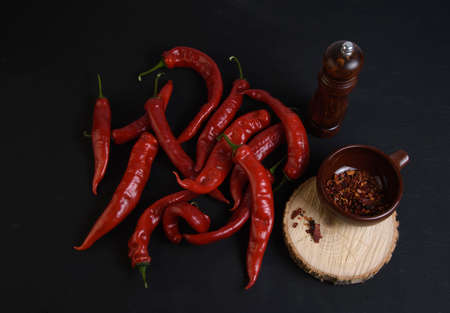 red bitter peppers on a black wooden table, next to a cut of a tree trunk, there is a plate with ground pepper and a wooden manual spice millの写真素材