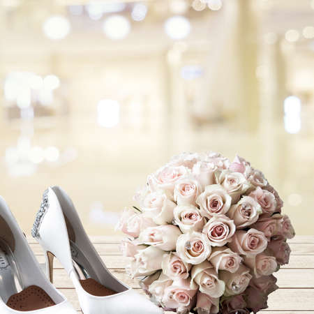 Glamorous white designer high heel shoes and a wedding bouquet with white and pink roses sitting on table with a wedding hall backgroundの写真素材
