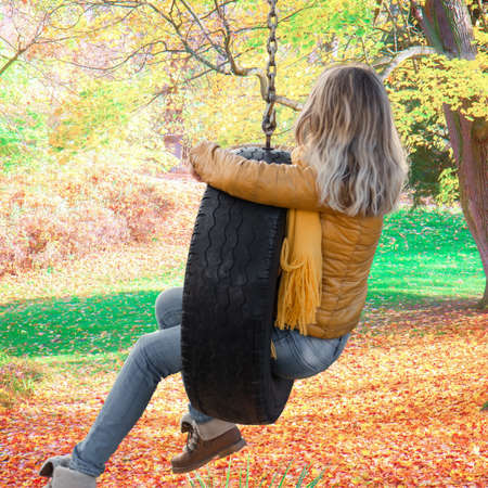 Young woman wearing a gold jacket sitting in a tire swing in the fall with leavesの写真素材