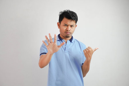 Attractive Asian man with hand gesture pose rejection or prohibition while pointing to copy space by his side wearing blue t shirt isolated on white backgroundの写真素材