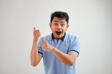 shocked asian man with an open mouth with fingers pointing to behind wearing blue t shirt isolated on white backgroundの写真素材