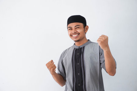 Successful happy young asian muslim man screaming shouting and shows winning victory gesture wearing grey muslim clothes isolated on white backgroundの写真素材