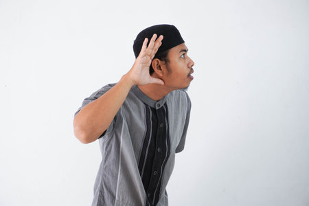 young Asian muslim man wearing grey muslim clothing, hands near ears trying to listening a gossip isolated on white backgroundの写真素材
