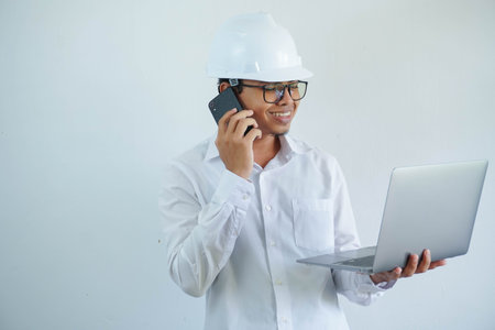 young Asian male engineer wearing white hard hat holding laptop and on call for construction work isolated on white background, copy space.の写真素材