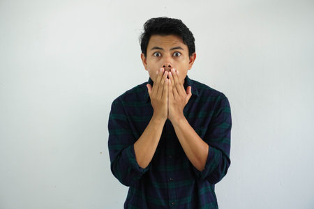 young asian man showing shocked expression with his hands covering mouth isolated on white...