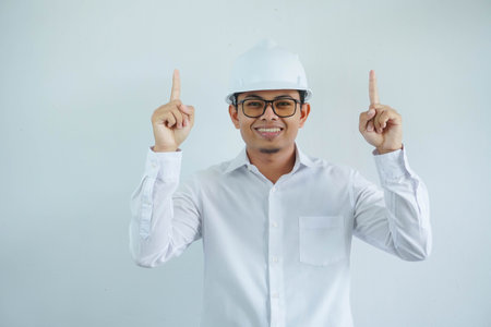 young asian architect man wearing white hard hat safety helmet looking camera with finger pointing up isolated on white backgroundの写真素材