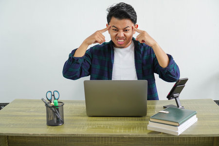 Frustrated young Asian businessman sitting at workplace because of piled up tasksの写真素材