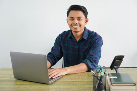 smiling young asian man in blue shirt sit work at wooden desk with pc laptop isolated on white...