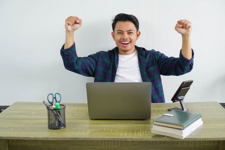 Young asian overjoyed fun employee business man in blue shirt sitting at workplace desk pc laptop do winner gesture clench fist isolated, Achievement career conceptの写真素材