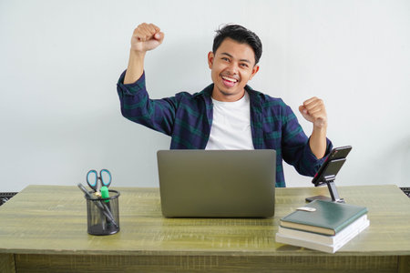 Young asian overjoyed fun employee business man in blue shirt sitting at workplace desk pc laptop do...