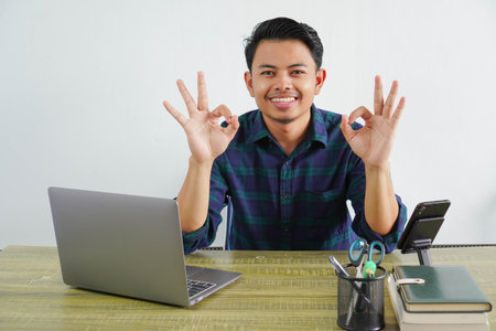 Smiling young asian man in blue shirt sit work at wooden desk with pc laptop isolated on white background. Achievement business career lifestyle concept. showing OK gestureの写真素材