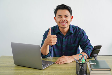 Smiling young asian man in blue shirt sit work at wooden desk with pc laptop isolated on white...