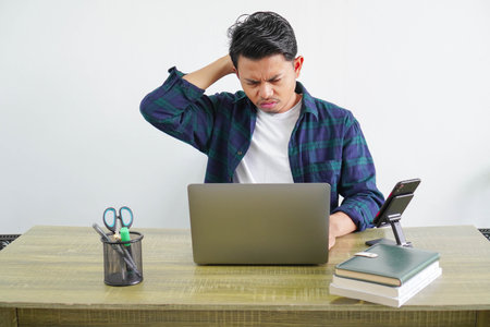 An Asian male freelance worker is sitting looking at a laptop showing a confused gesture with his...