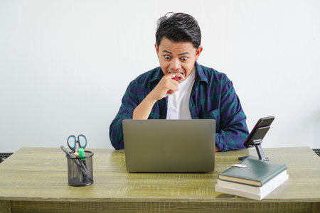 worried asian man in a workplace looking at laptop screen while biting finger