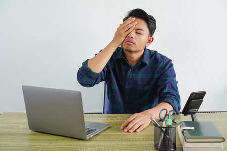 young asian man in blue shirt sit work at wooden desk with pc laptop isolated on white background. Achievement business career lifestyle concept. put palm on face, keeping eyes closedの写真素材