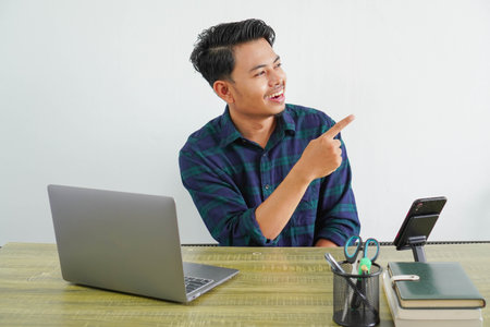 smiling young asian man sit work at wooden desk with pc laptop. Achievement business career lifestyle concept. pointing index finger aside upの写真素材