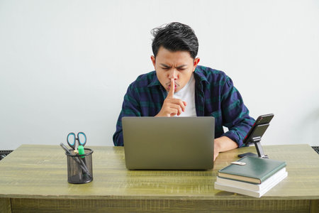 An Asian male freelance worker is sitting showing a silent gesture with his finger touching his lipsの写真素材
